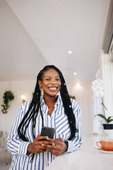 Cheerful young businesswoman smiling at the camera while holding a smartphone in a coffee shop. Happy young black businesswoman enjoying her coffee break in a cafe. - JLPSF28595