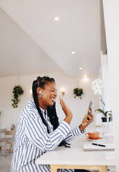 Glückliche junge Geschäftsfrau winkt bei einem Videoanruf, während sie in einem Café arbeitet. Lächelnde schwarze Geschäftsfrau im Video-Chat mit einem ihrer Kunden in einem Café. - JLPSF28600