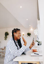 Happy young businesswoman waving a greeting on a video call while working in a cafe. Smiling black businesswoman video chatting with one of her clients in a coffee shop. - JLPSF28600