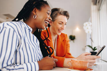 Zwei Kolleginnen lächeln fröhlich, während sie in einem Bürocafé gemeinsam ein Smartphone benutzen. Glückliche Geschäftsfrauen, die ihre Kaffeepause an einem modernen Arbeitsplatz genießen. - JLPSF28602