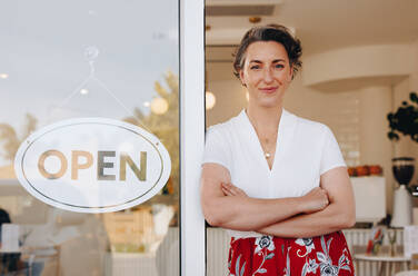Reife Kleinunternehmerin, die in die Kamera schaut, während sie neben einem offenen Schild am Eingang ihres Cafés steht. Unternehmerin, die Kunden in ihrem neu eröffneten Café willkommen heißt. - JLPSF28605
