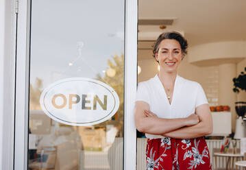 Erfolgreiche Café-Besitzerin lächelt in die Kamera, während sie neben einem offenen Schild am Eingang ihres Restaurants steht. Eine Kleinunternehmerin, die Kunden in ihrem neu eröffneten Café willkommen heißt - JLPSF28606