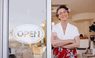 Glückliche Coffeeshop-Besitzerin lächelt in die Kamera, während sie neben einem offenen Schild am Eingang ihres Cafés steht. Reife Kleinunternehmerin, die Kunden in ihrem neu eröffneten Restaurant willkommen heißt. - JLPSF28608