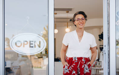 Reife Frau lächelt in die Kamera, während sie neben einem offenen Schild am Eingang ihres Restaurants steht. Glückliche Kleinunternehmerin, die Kunden in ihrem neuen Café willkommen heißt. - JLPSF28609