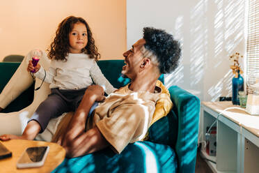 Entzückendes kleines Mädchen, das in die Kamera schaut, während es auf dem Sofa auf seinem Vater sitzt. Glücklicher Vater und Tochter, die zusammen zu Hause Spaß haben. Vater und Kind, die eine schöne Zeit miteinander verbringen. - JLPSF28612