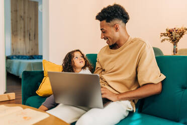 Glücklicher alleinerziehender Vater, der seine Tochter anlächelt, während er mit einem Laptop auf einer Couch sitzt. Liebevoller Vater, der seine Tochter mit Hilfe von Online-Bildungsinhalten zu Hause unterrichtet. Vater und Tochter sehen sich Lernvideos an. - JLPSF28615