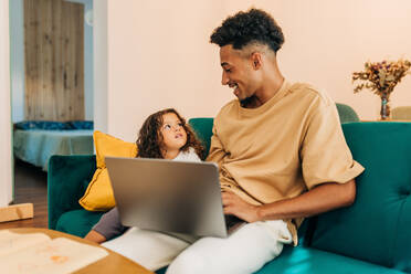 Happy single dad smiling at his daughter while sitting on a couch with a laptop. Loving dad homeschooling his daughter using online educational content. Father and daughter watching tutorial videos. - JLPSF28615