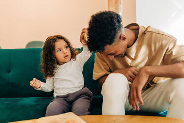 Niedliches kleines Mädchen kämmt ihrem Vater die Haare, während sie auf einer Couch im Wohnzimmer sitzt. Liebenswerter Vater und Tochter, die zu Hause verspielt sind. Vater und Tochter verbringen Zeit miteinander. - JLPSF28619