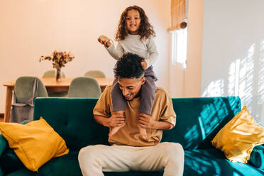 Adorable little girl smiling at the camera while sitting on her father's shoulders. Happy little girl having fun with her dad at home. Father and daughter spending some quality time together. - JLPSF28621