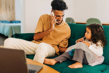 Smiling single father looking at his daughter while speaking on a phone call. Happy young father sitting on a couch with his little daughter. Father spending time with his daughter at home. - JLPSF28628