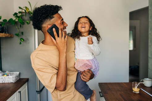 Fröhlicher alleinstehender Vater, der lacht und telefoniert, während er seine Tochter bei sich trägt. Glücklicher alleinstehender Vater, der seine Zeit zu Hause mit seiner bezaubernden Tochter verbringt. - JLPSF28634