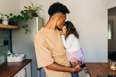 Liebevoller Vater, der seiner Tochter einen Kuss auf die Stirn gibt, während er sie in seinen Armen trägt. Liebevoller alleinerziehender Vater, der sein kleines Mädchen zu Hause tröstet. - JLPSF28636