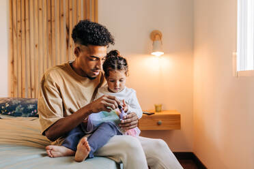 Junger Vater, der die Nägel seiner Tochter mit Nagellack lackiert, während er auf einem Bett sitzt. Junger alleinerziehender Vater, der einige Zeit mit seiner bezaubernden Tochter zu Hause verbringt. - JLPSF28640