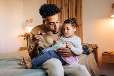 Alleinstehender Vater bläst Nagellack auf die Nägel seiner Tochter. Junger Vater lackiert die Nägel seiner Tochter, während er auf einem Bett sitzt. Liebenswerter Vater und Tochter verbringen einige Zeit zu Hause. - JLPSF28642