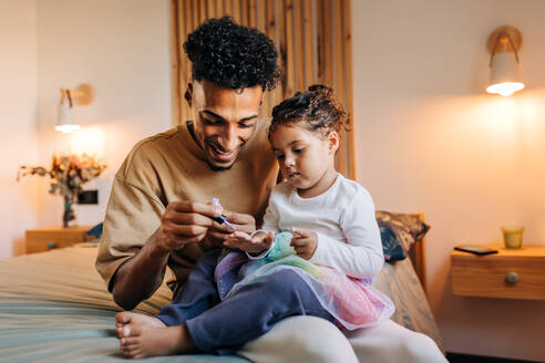 Lächelnder alleinerziehender Vater, der die Nägel seiner Tochter mit Nagellack lackiert, während er auf einem Bett sitzt. Glücklicher junger Vater, der einige Zeit mit seiner bezaubernden Tochter zu Hause verbringt. - JLPSF28643