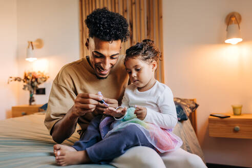 Smiling single dad painting his daughter's nails with nail polish while sitting on a bed. Happy young father spending some quality time with his adorable daughter at home. - JLPSF28643