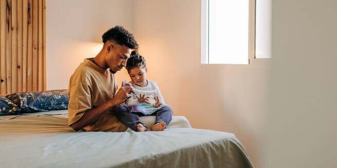 Vater trägt Nagellack auf die Nägel seiner Tochter auf, während er zu Hause auf dem Bett sitzt. Moderner alleinerziehender Vater, der einige Zeit mit seiner bezaubernden Tochter in einem Haus verbringt. - JLPSF28644