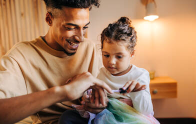 Adorable little girl applying nail polish on her dad's nails. Playful father and daughter spending some quality time together at home. - JLPSF28645