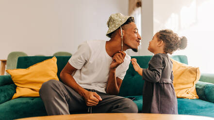 Verspielter Vater, der seiner Tochter einen Kuss gibt, während er auf der Couch im Wohnzimmer sitzt. Vater, der sich mit seiner kleinen Tochter zu Hause amüsiert. Vater und Tochter verbringen eine schöne Zeit miteinander. - JLPSF28648