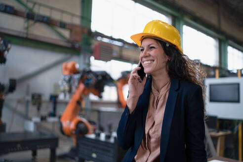 Eine Automatisierungsingenieurin mit einem Tablet zur Programmierung eines Roboterarms telefoniert mit ihrem Mobiltelefon und geht durch die Fabrik. - HPIF00708