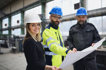 A manager supervisors, engineer and industrial worker in uniform discussing blueprints in large metal factory hall. - HPIF00768