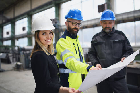 Ein leitender Angestellter, ein Ingenieur und ein Industriearbeiter in Uniform besprechen Blaupausen in einer großen Metallfabrikhalle. - HPIF00768