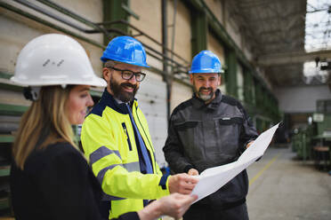 Ein leitender Angestellter, ein Ingenieur und ein Industriearbeiter in Uniform besprechen Blaupausen in einer großen Metallfabrikhalle. - HPIF00769