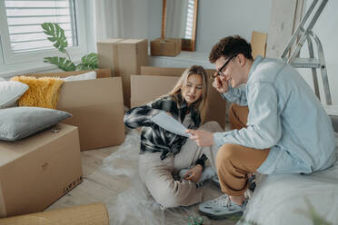 A cheerful young couple in their new apartment, sitting on floor and reading papers. Conception of moving. - HPIF00793