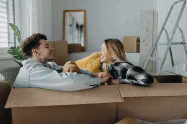 Ein fröhliches junges Paar in seiner neuen Wohnung, das in Kartons sitzt und sich amüsiert. Konzept des Umzugs. - HPIF00801