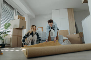 A cheerful young couple in their new apartment, rolling out carpet. Conception of moving. - HPIF00811