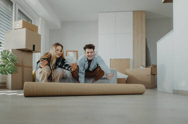 Ein fröhliches junges Paar in ihrer neuen Wohnung, das einen Teppich ausrollt. Vorstellung eines Umzugs. - HPIF00812