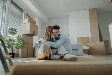 Ein fröhliches junges Paar in seiner neuen Wohnung, das auf dem Boden sitzt und sich umarmt, Vorstellung eines Umzugs. - HPIF00813