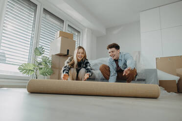 A cheerful young couple in their new apartment, rolling out carpet. Conception of moving. - HPIF00814