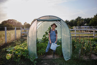 A senior gardener woman holding watering can in greenhouse at garden, looking at camera. - HPIF00884
