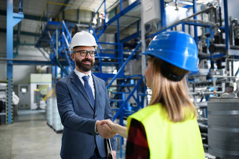 Ein Ingenieur und ein Industriearbeiter in Uniform geben sich in einer großen Metallfabrikhalle die Hand und unterhalten sich. - HPIF00904