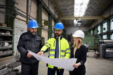 Ein leitender Angestellter, ein Ingenieur und ein Industriearbeiter in Uniform besprechen Blaupausen in einer großen Metallfabrikhalle. - HPIF00911