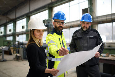 Ein leitender Angestellter, ein Ingenieur und ein Industriearbeiter in Uniform besprechen Blaupausen in einer großen Metallfabrikhalle. - HPIF00912