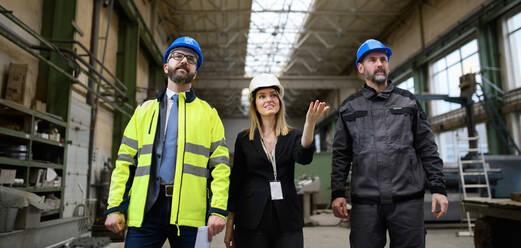 Manager supervisors and industrial worker in a uniform walking in large metal factory hall and talking. Low angle view. - HPIF00913