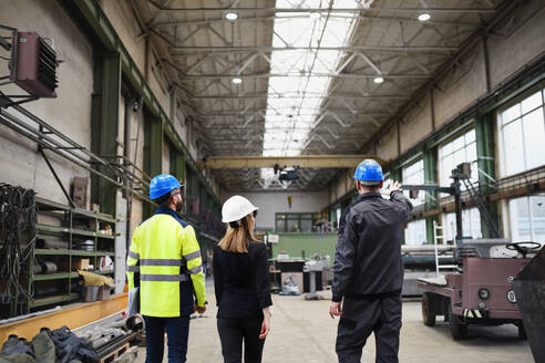Eine Rückansicht von leitenden Angestellten und Industriearbeitern in Uniform, die in einer großen Metallfabrikhalle spazieren gehen und sich unterhalten. - HPIF00914