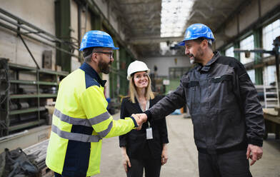 Ein Ingenieur und ein Industriearbeiter in Uniform geben sich in einer großen Metallfabrikhalle die Hand und unterhalten sich. - HPIF00916