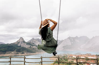 Unrecognizable brunette in romantic dress and sunhat sitting on swing near calm lake and small town against mountain range and cloudy gray sky in Riano in Leon, Spain - ADSF40380