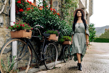 Ganzkörper einer lächelnden Frau in einem Kleid mit Sonnenhut, die lächelnd nach oben schaut, während sie auf dem Bürgersteig in der Nähe von Fahrrädern und einer Steinmauer in der Stadt spazieren geht - ADSF40388