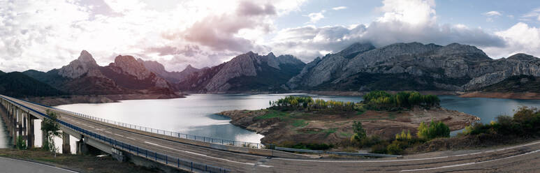 Panoramic view of long bridge over lake against mountains and green trees on coast under cloudy sky - ADSF40389