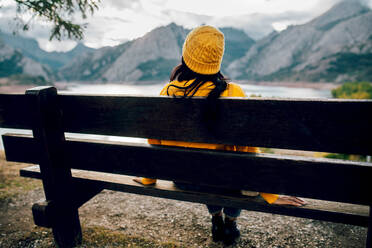 Rückenansicht einer anonymen Brünetten in orangefarbenem Pullover und Mütze, die auf einer Holzbank sitzt und die wunderbare Landschaft eines Bergkamms und eines ruhigen Sees genießt - ADSF40390