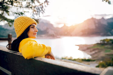 Seitenansicht Frau in orangefarbenen Pullover und Mütze sitzen auf Holzbank und genießen erstaunliche Landschaft der Bergkamm und ruhigen See - ADSF40394