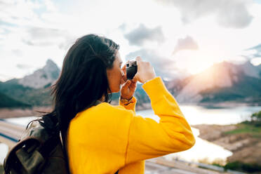 Seitenansicht einer weiblichen Reisenden in orangefarbenem Pullover mit Rucksack, die in der Nähe eines Sees steht und mit einer Fotokamera ein Bild von den Bergen macht - ADSF40397