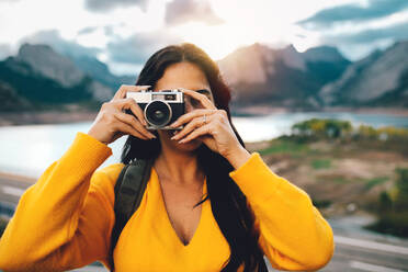 Unerkennbare weibliche Touristin, die ein Foto mit einer alten Fotokamera macht, während sie auf einer Straße in der Nähe eines Sees gegen Berge unter bewölktem Himmel steht - ADSF40399