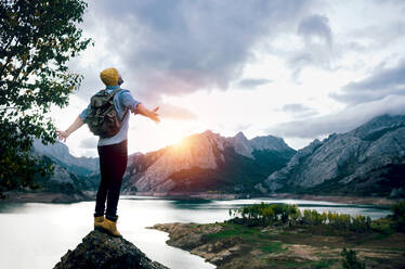 Unrecognizable backpacker standing on rocky peak and admiring picturesque scenery of mountains and lake under cloudy sky in daylight - ADSF40401