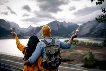 Back view of backpacker and girlfriend standing with burning sparklers against mountain ridge while traveling together in cloudy day - ADSF40402