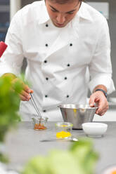Aufmerksamer Mann in weißer Kochjacke, der ein Gericht in einer Edelstahlschüssel zubereitet und ein Metallwerkzeug in einer Restaurantküche benutzt - ADSF40412